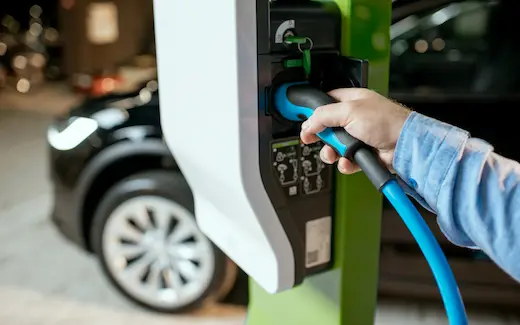 A greyscale photo of a hand plugging a cable for an electric car into the electric vehicle charging station