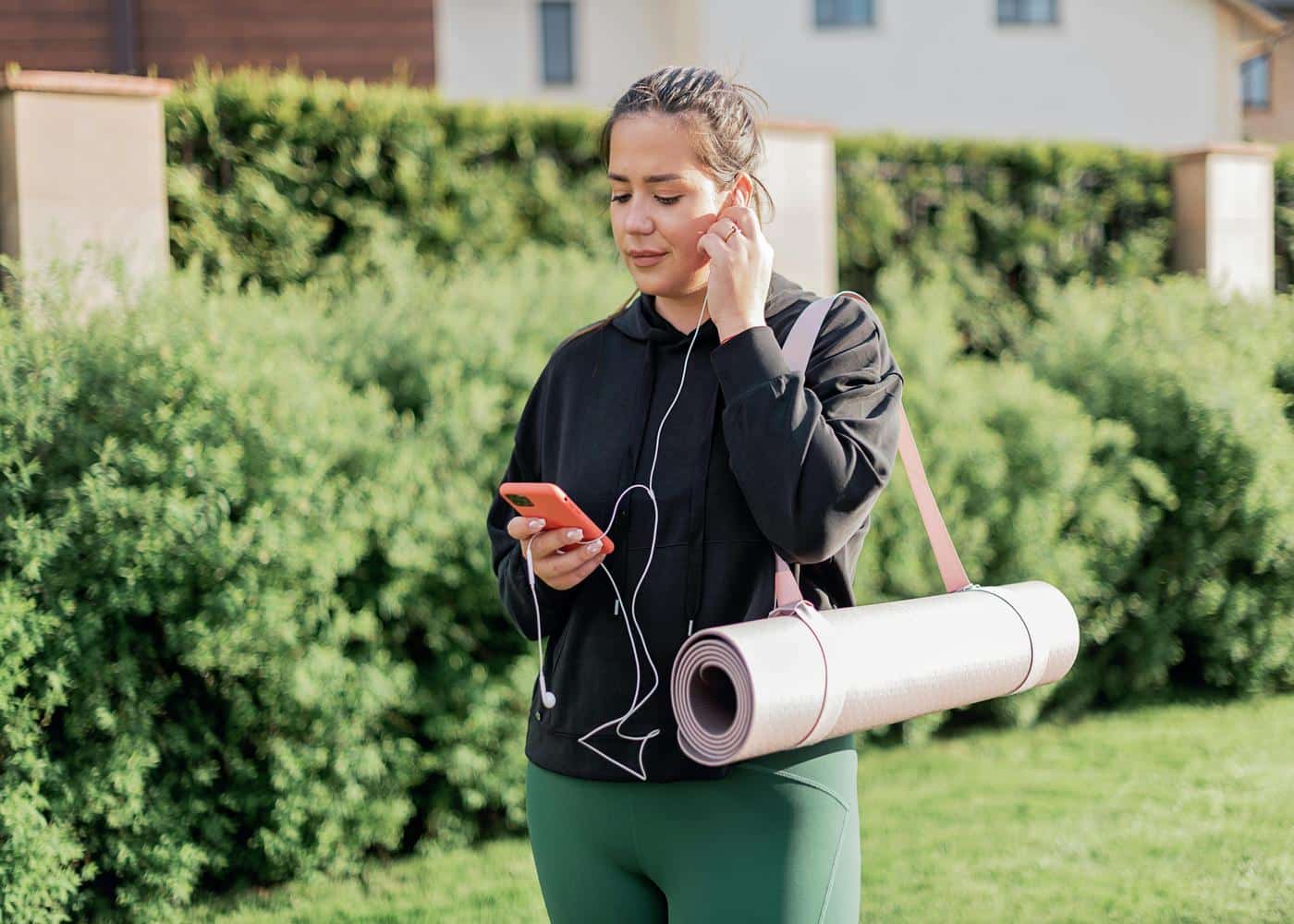 Woman in workout clothes with strapped yoga mat around one arm and headphones on looking at smartphone