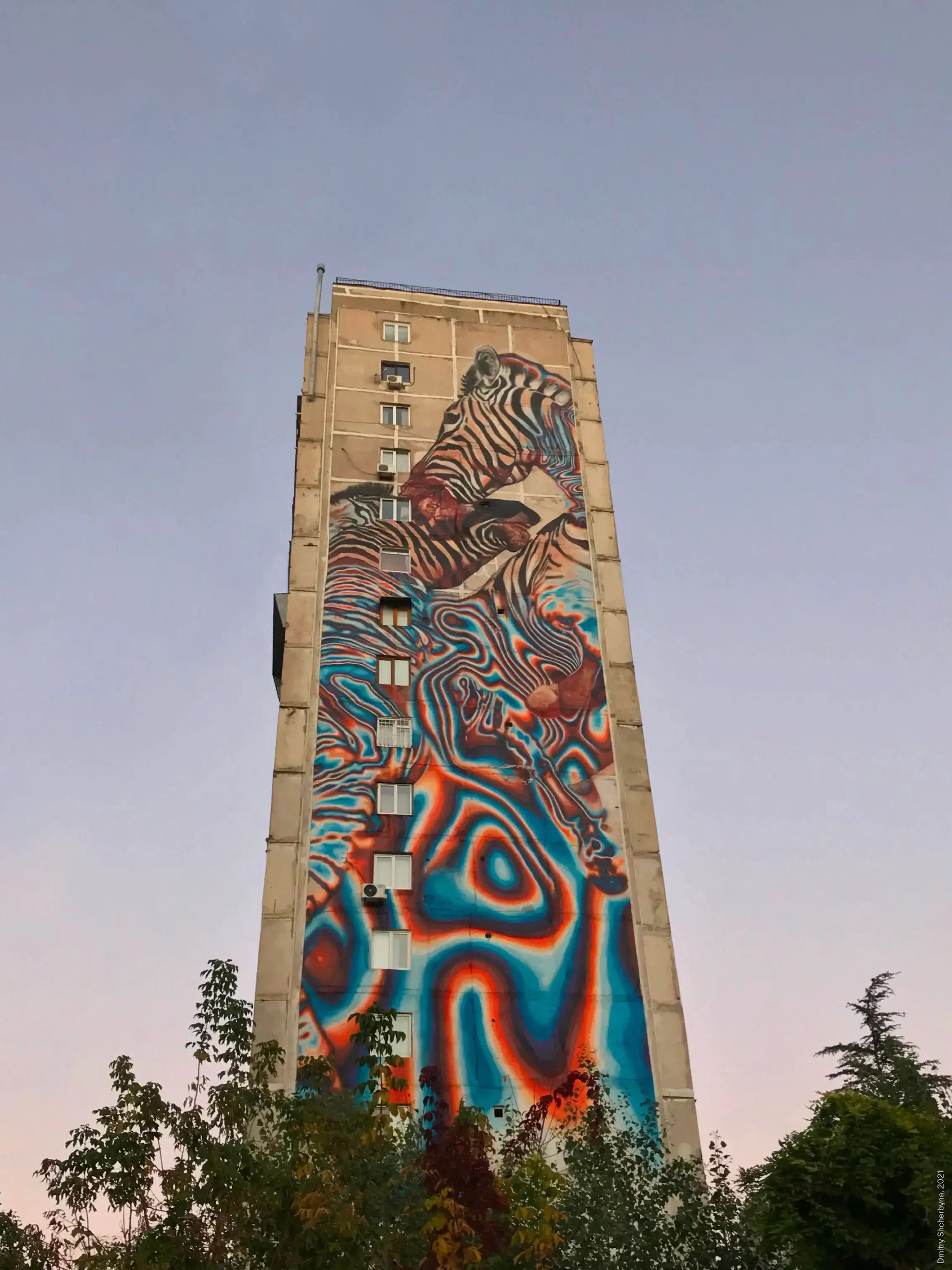A mural covering an entire apartment building depicting zebras that dissolve into swirling psychedelic patterns in cyan blue, orange and red.