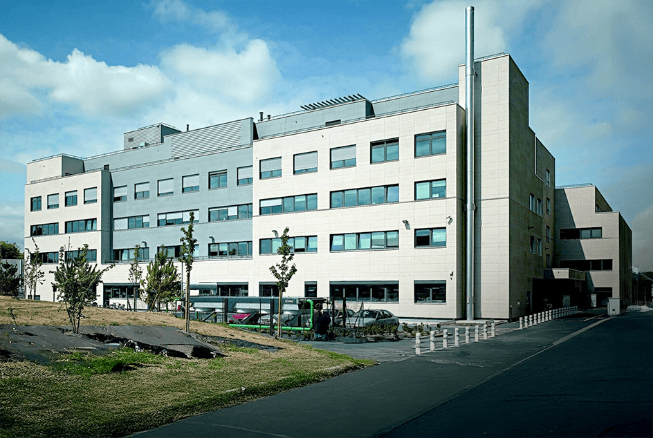 Modern multi-story hospital building exterior with clean architectural design, featuring multiple floors with windows and a contemporary facade under a blue sky with clouds