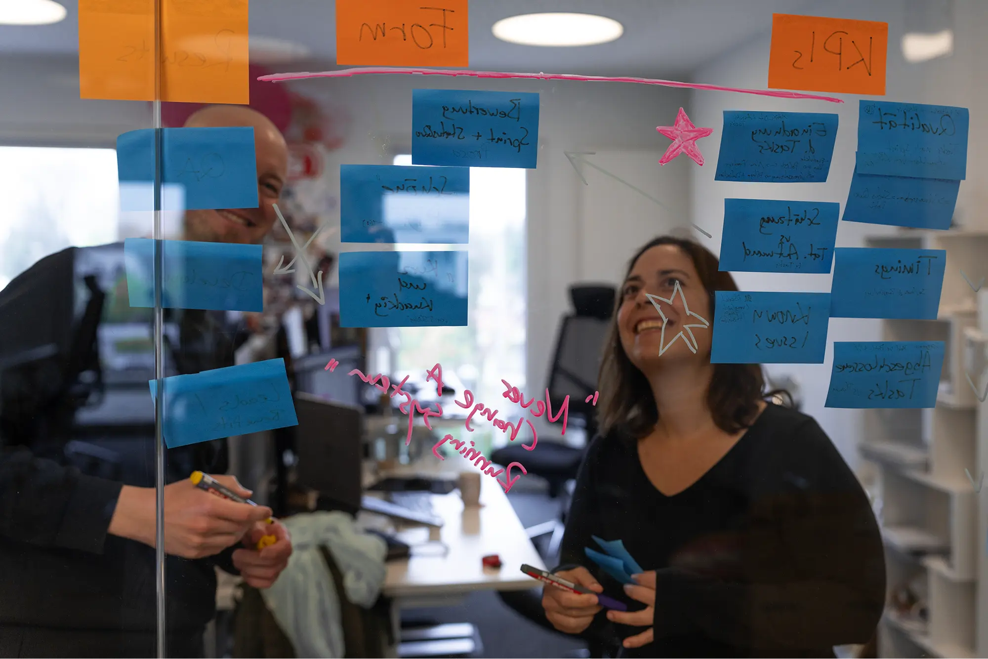 Ein Teammitglied und eine Kollegin arbeiten gut gelaunt im Büro zusammen vor einer gläsernen Trennwand, die mit blauen und orangefarbenen Post-its, Skizzen und handschriftlichen Projektnotizen beklebt ist.