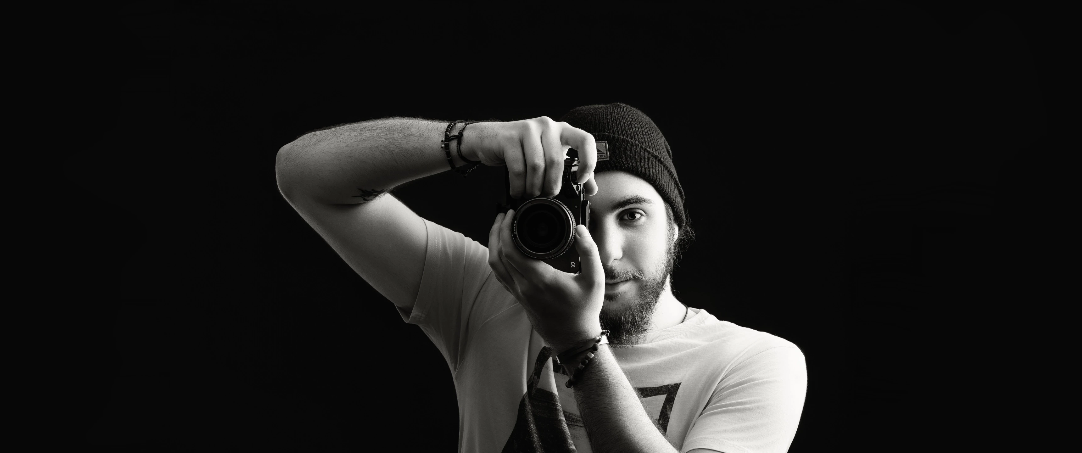 Black and white portrait of videographer holding a camera to eye, capturing a shot against a dark background, representing Raybrand Media’s professional videography, creative storytelling and visual content production