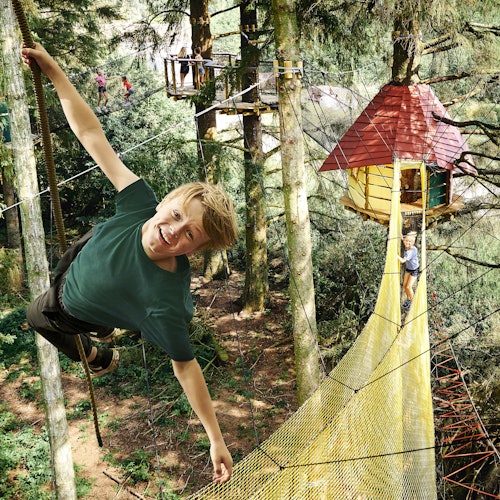 森のアドベンチャーパークで微笑む少年がロープにぶら下がっています。背景には、黄色いネットの橋を渡るとツリーハウスがあります。