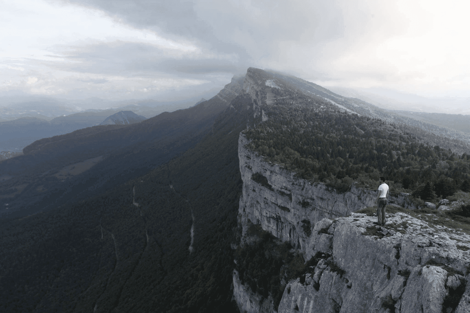 Man standing on the edge of a dramatic limestone cliff, overlooking misty mountain ridges in the French Alps — a moment of solitude and awe.