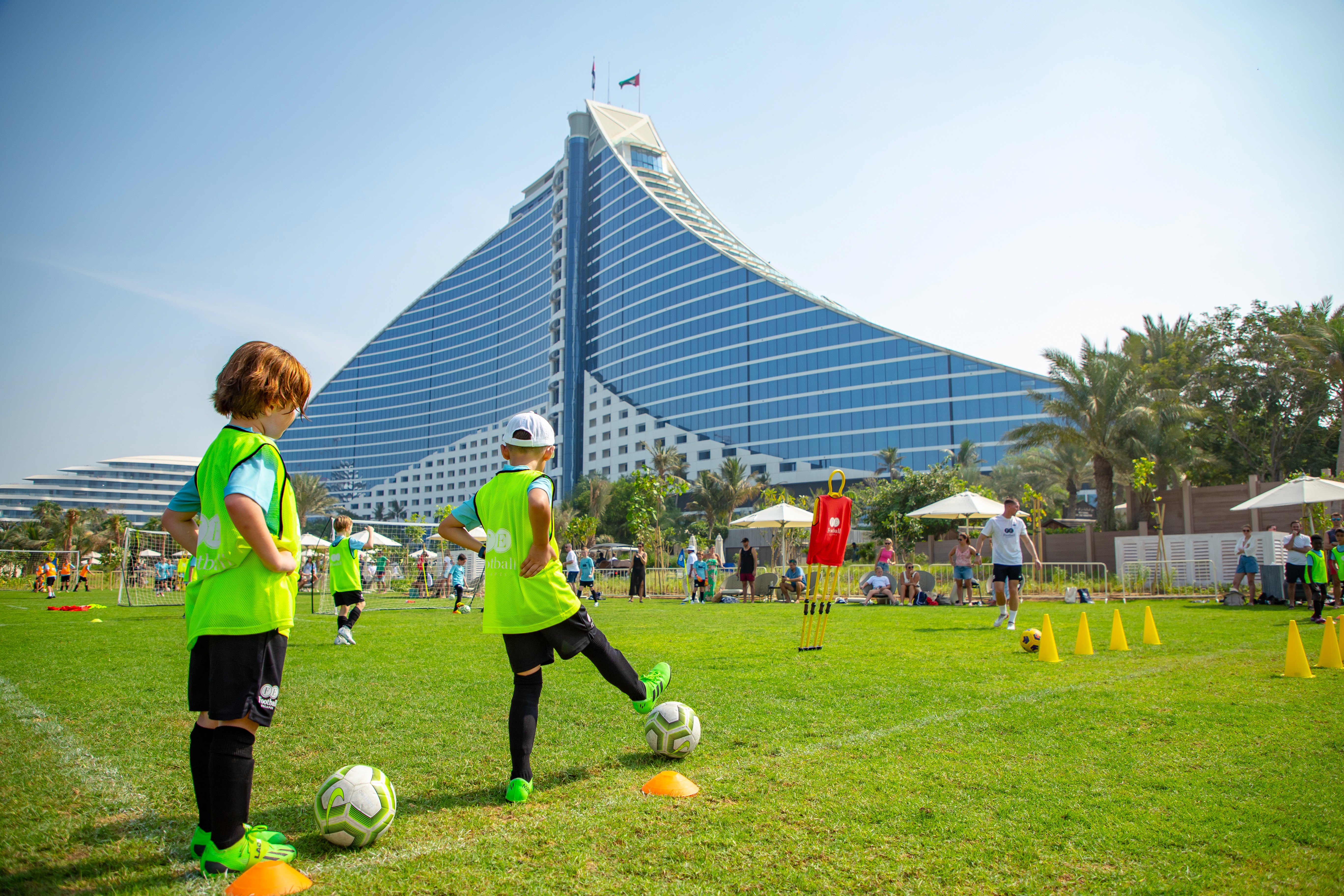 Children practicing football drills on a grass field filled with obstacles. The Jumeirah Beach Hotel is in the background with the clear, blue sky.