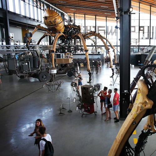 Pessoas dentro de um grande armazém moderno com uma aranha robótica gigante e várias exposições mecânicas.