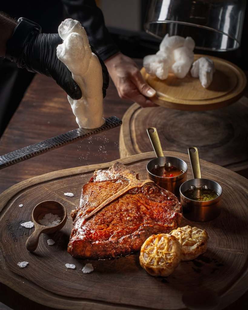 A beautifully cooked steak seasoned with salt, served on a wooden platter alongside two small bowls of sauces, garnished with grilled lemon slices.