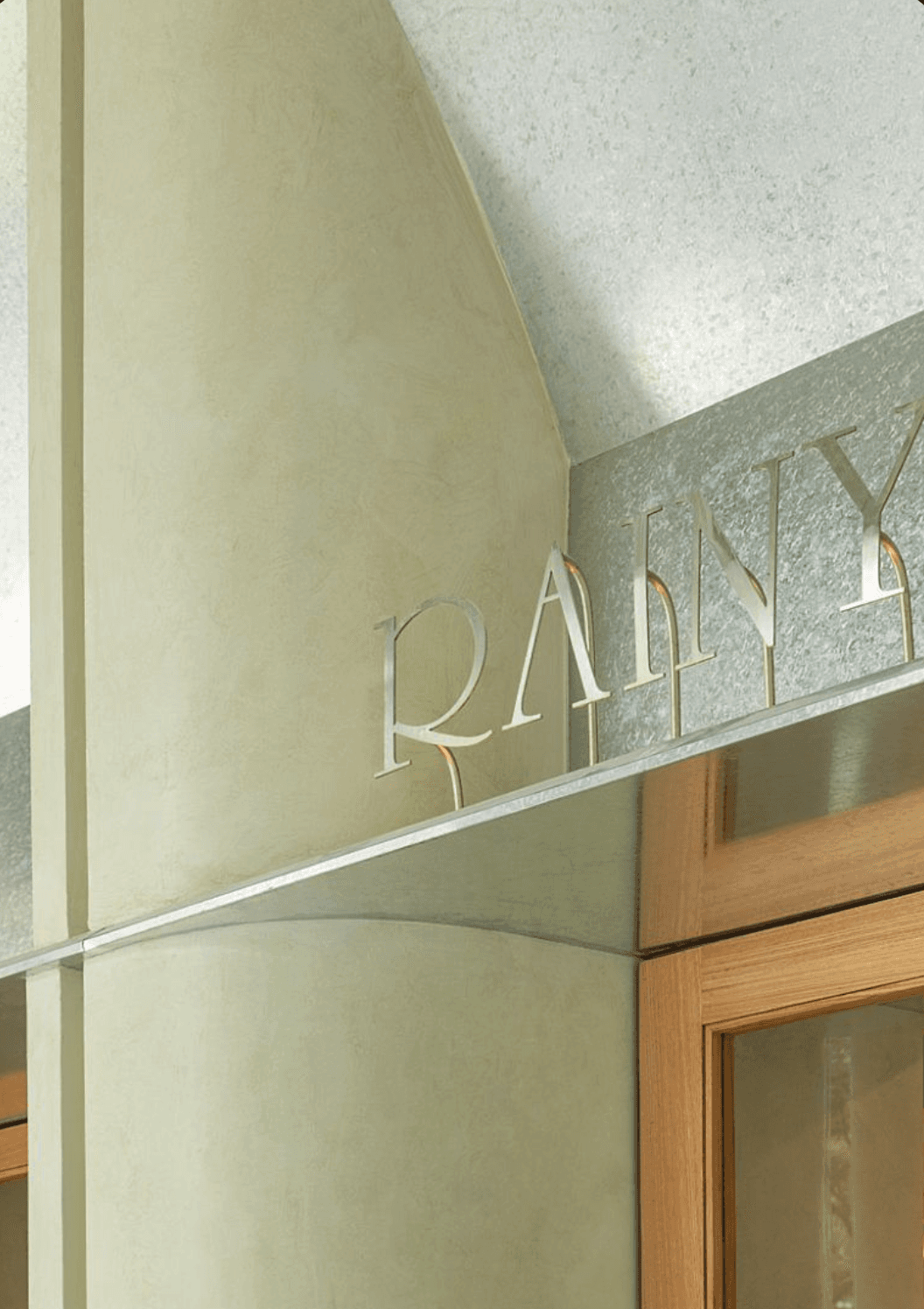 Close-up of polished brushed steel dimensional metal letters spelling 'RAINY' in an elegant serif typeface, mounted along a textured sage green stone fascia above a timber-framed glass entrance — letters catch the light against the muted architectural surface.