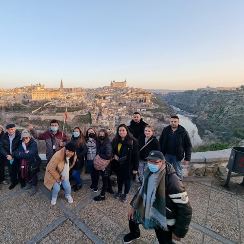 A group of people stands on a viewpoint overlooking a historic cityscape with a river running through a valley below.