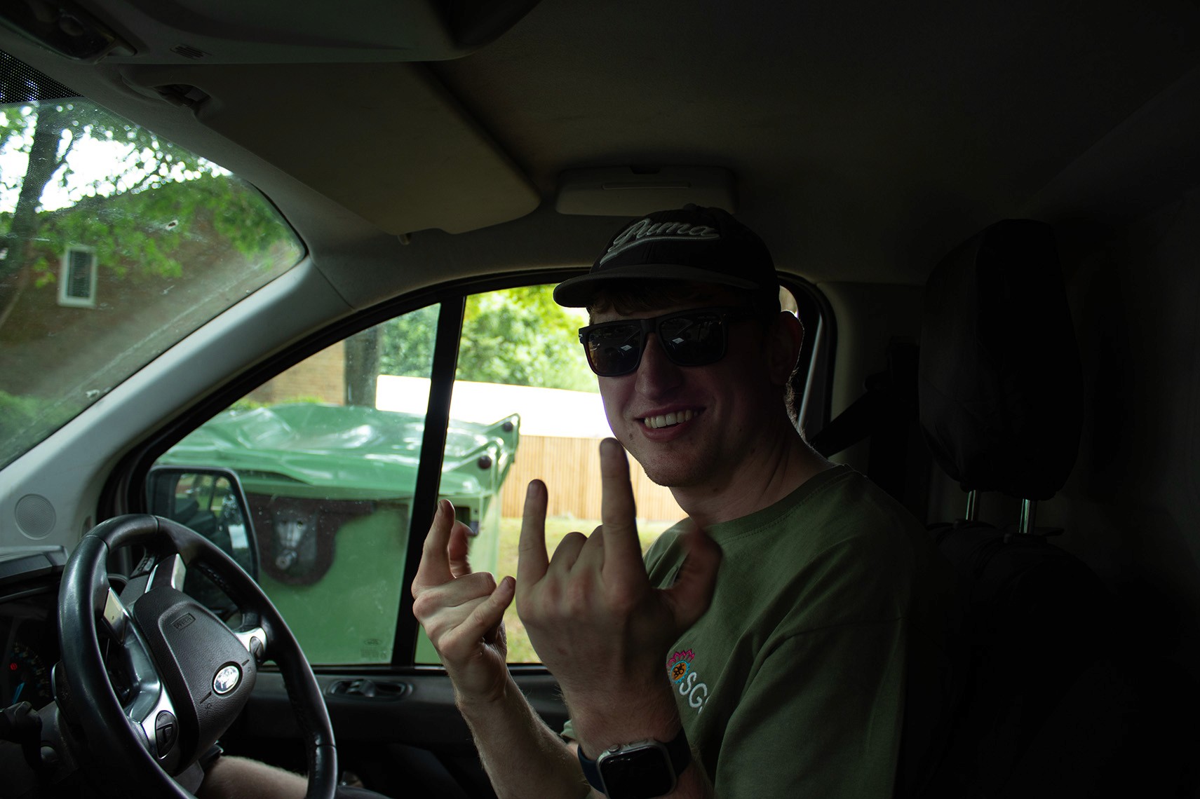 A person sitting in a vehicle, making a hand gesture while looking towards the camera.