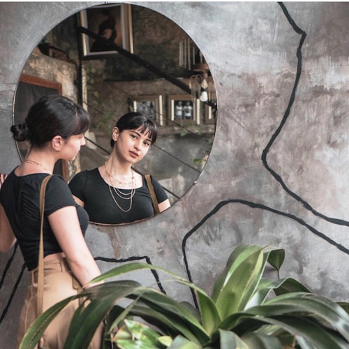 一位女性在一面安裝在質感牆壁上的圓形鏡子中注視著自己的倒影，前景中有一株植物。