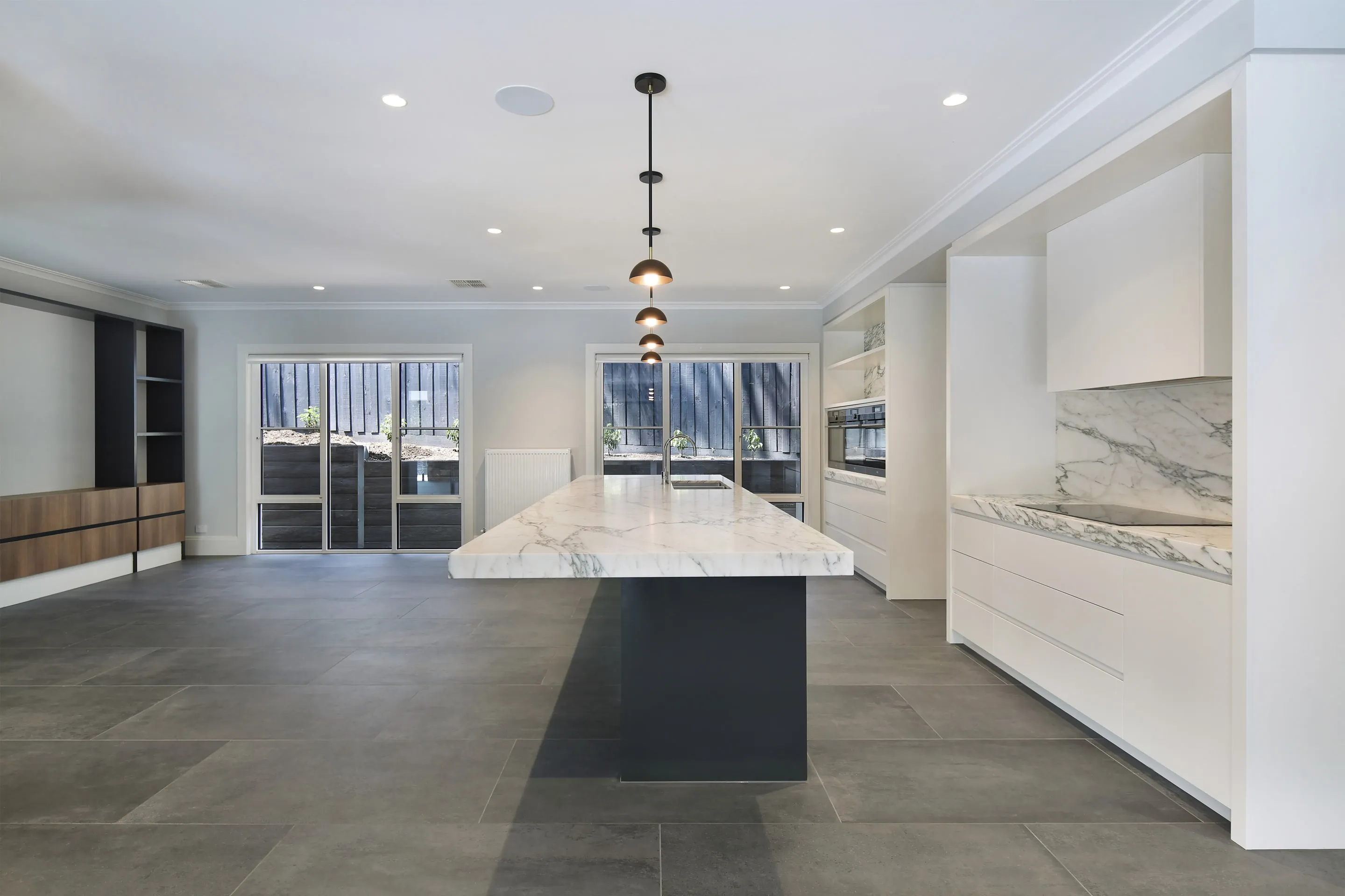 Kitchen and living zone with a marble waterfall island on a charcoal base, copper dome pendants, white joinery, marble splashback, a timber media wall and charcoal stone floors
