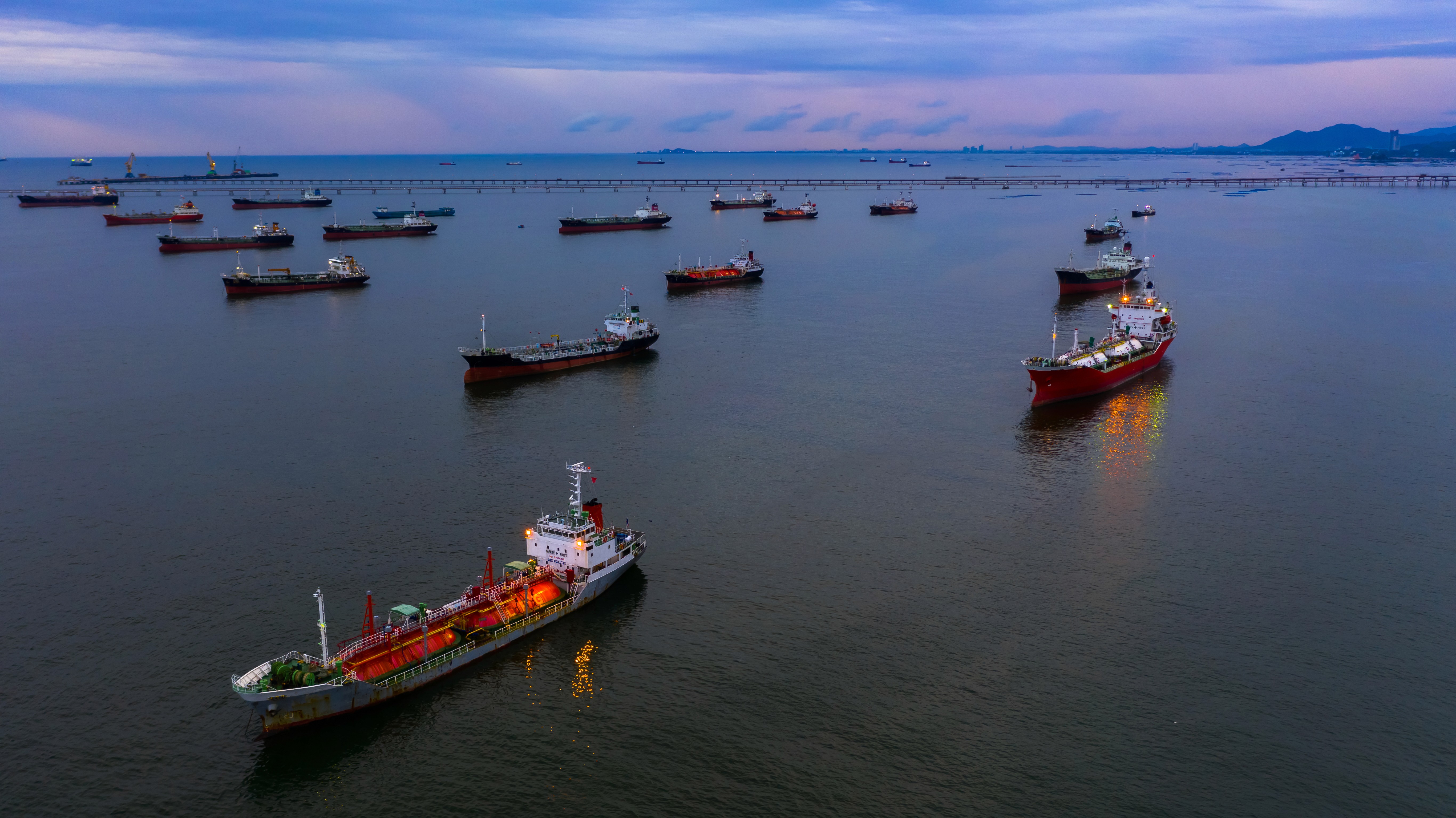 Aerial view of oil and gas tankers at anchor in calm water at dusk, with a long jetty on the horizon