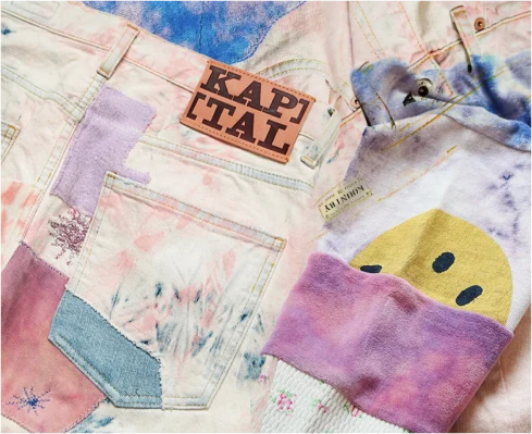 A selection of pastel-colored, tie-dye patterned clothing items, including a pair of jeans and a sweatshirt with playful details and the brand tag "KAPITAL."