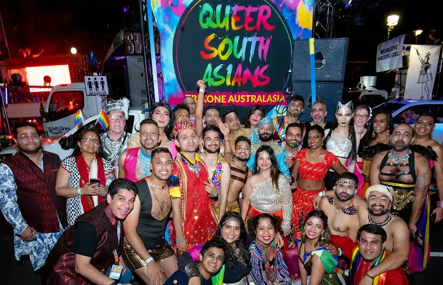 A large, diverse group of people in colourful, festive attire pose together, smiling, in front of a vibrant sign that reads "Queer South Asians, Trikone Australasia" at a lively event or parade.