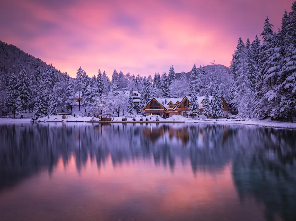 Winter sunset at Lake Jasna, Kranjska Gora, with snow-covered pine trees and a mountain lodge reflected in the calm lake water.
