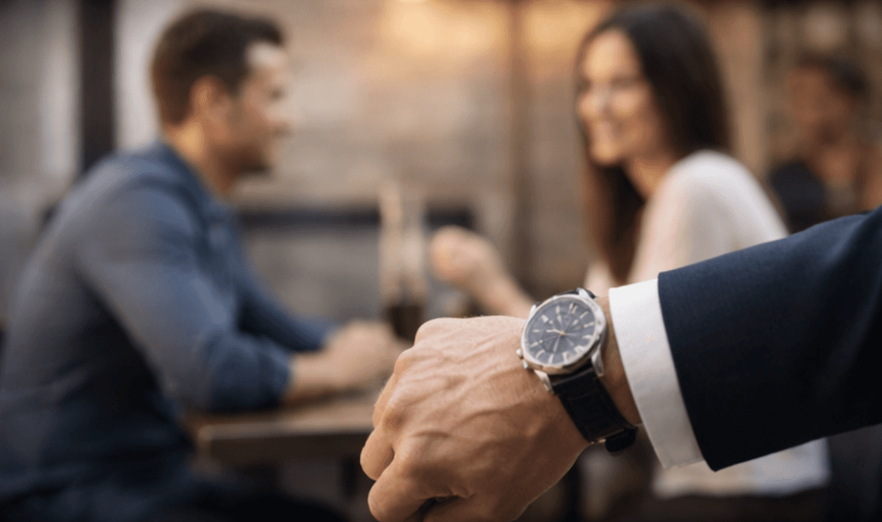 ein anzug tragender arm mit einer uhr ist im vordergrund, im hintergrund ist ein paar, das an einem tisch sitzt