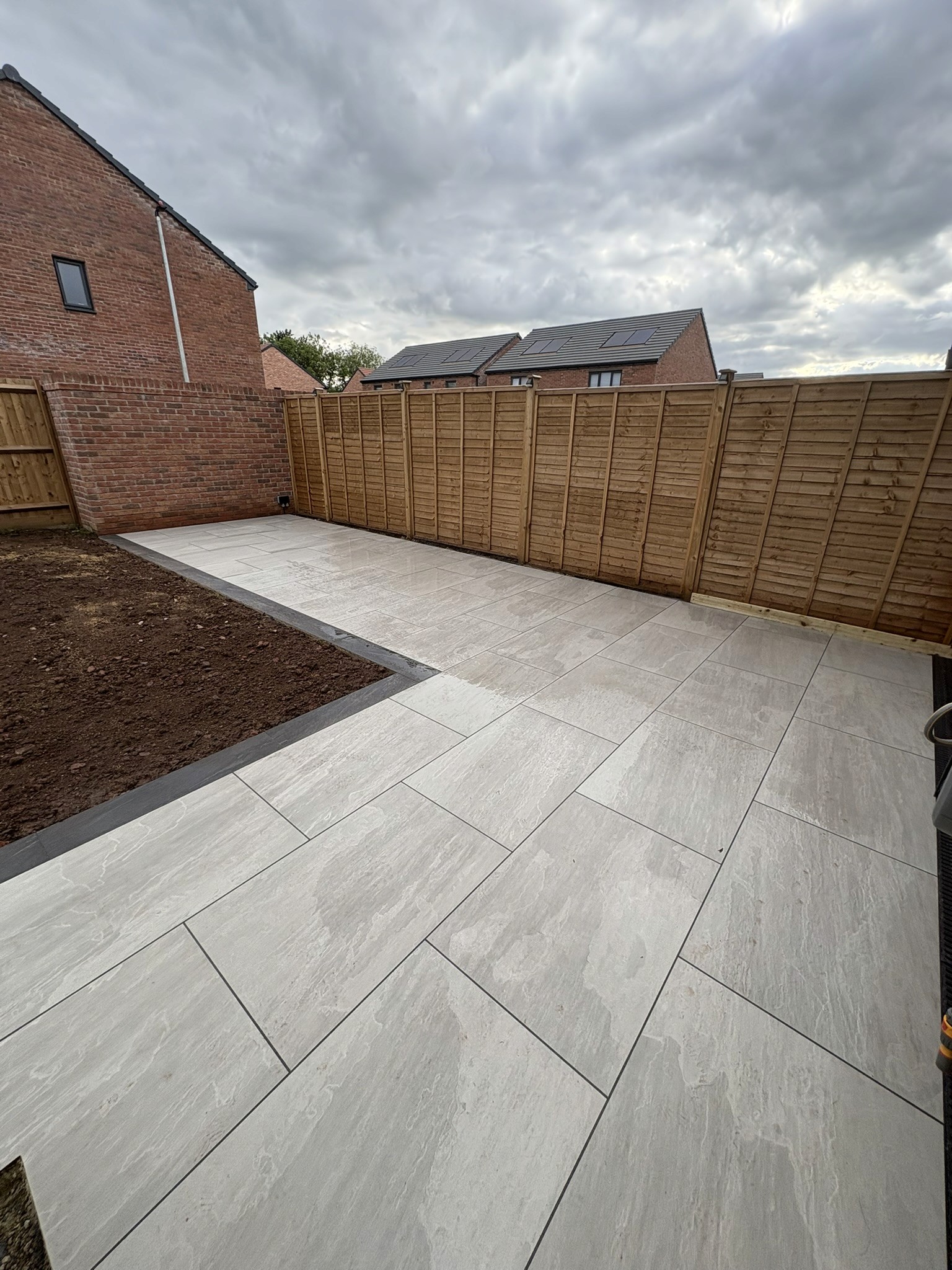 Porcelain patio beside a brick house, with timber fencing and an adjoining soil border.