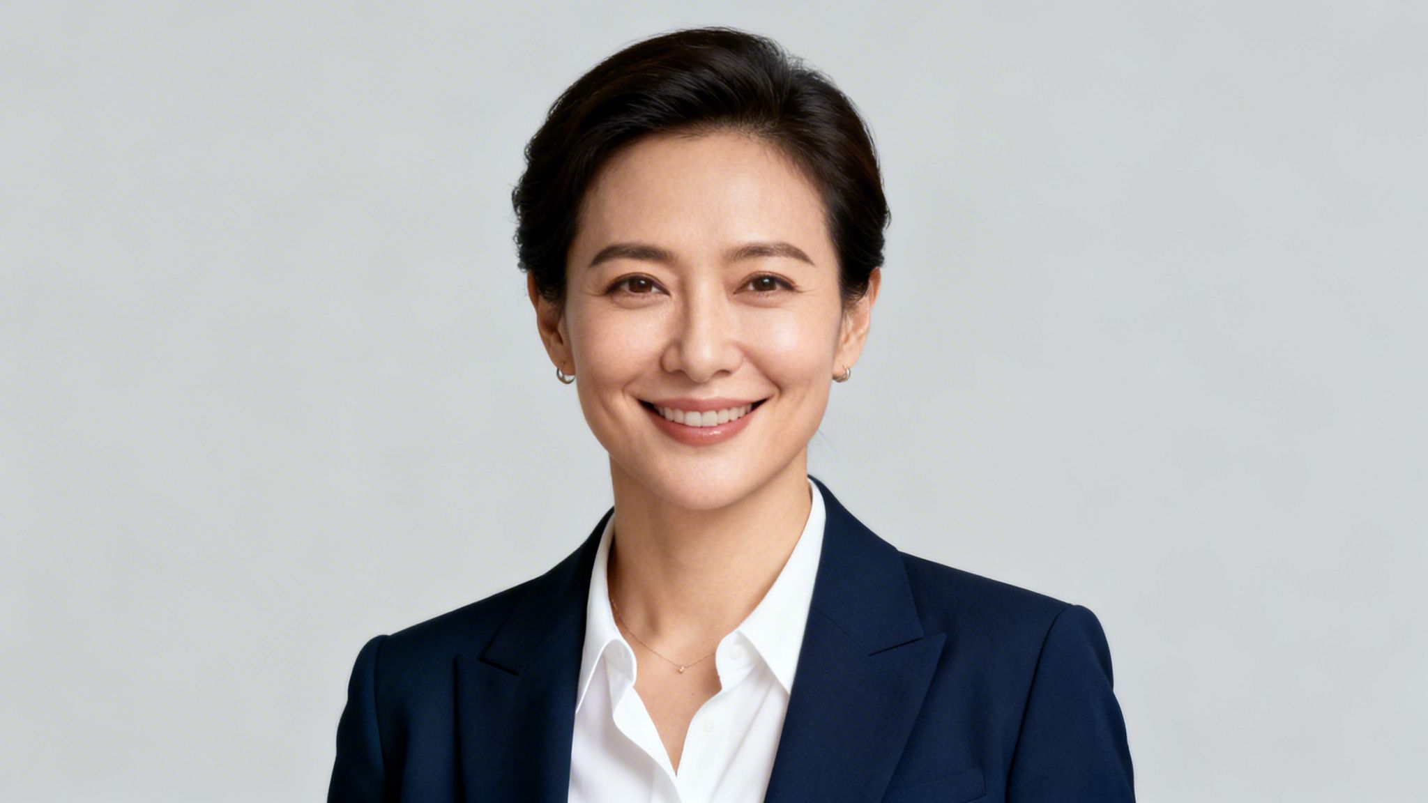Porträt einer lächelnden Geschäftsfrau mit kurzen dunklen Haaren in einem dunkelblauen Blazer und weißem Hemd vor grauem Hintergrund.