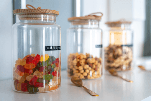 Drei Glasbehälter mit Holzdeckeln auf weißer Oberfläche, im Vordergrund ein Glas mit bunten Gummibärchen, dahinter zwei Gläser mit Nüssen und Trockenfrüchten, davor ein goldfarbenes Löffelchen