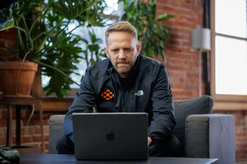 Close-up portrait of a person wearing a gray insulated hooded jacket with the hood up, showing their face framed by the hood against a light background.