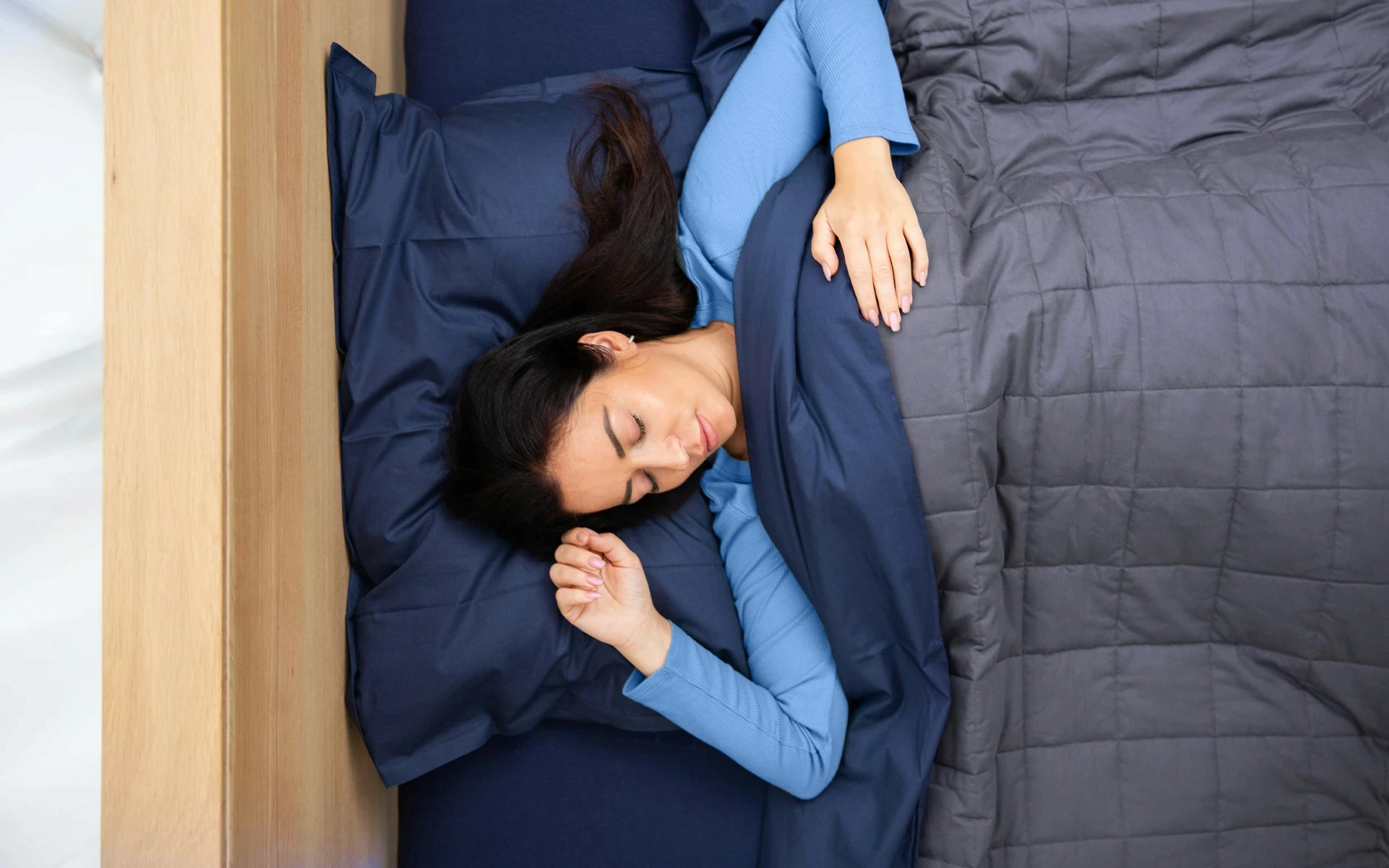 Woman sleeping peacefully on her side under dark blue bedding, resting on a navy pillow with a grey quilt.