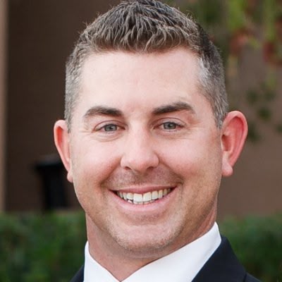 A smiling man in a suit stands outdoors, showing a bright demeanor against a blurred natural background.