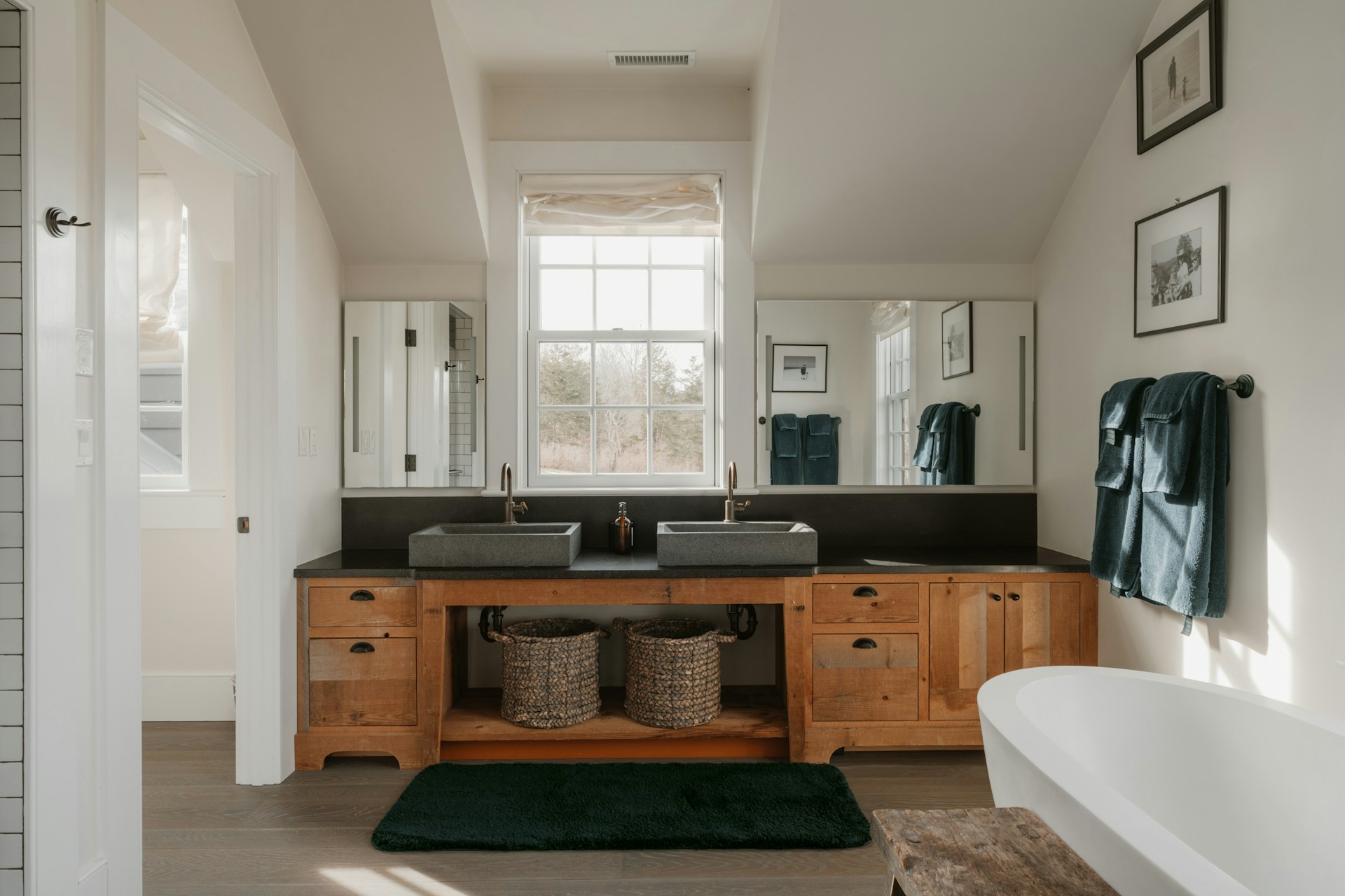 Bright rustic bathroom with a wood double vanity, stone sinks, a large window, dark green rug, and a white soaking tub.