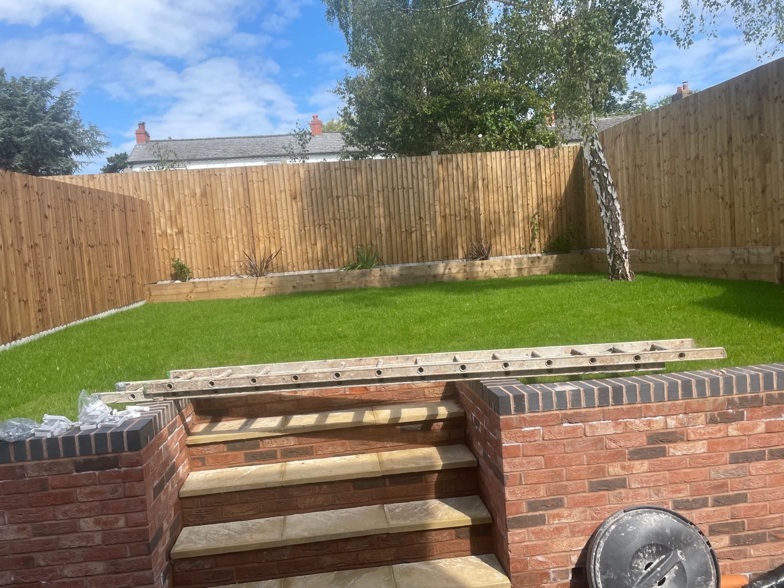 A well-maintained garden with green grass, brick steps, and wooden fences under a blue sky with clouds.