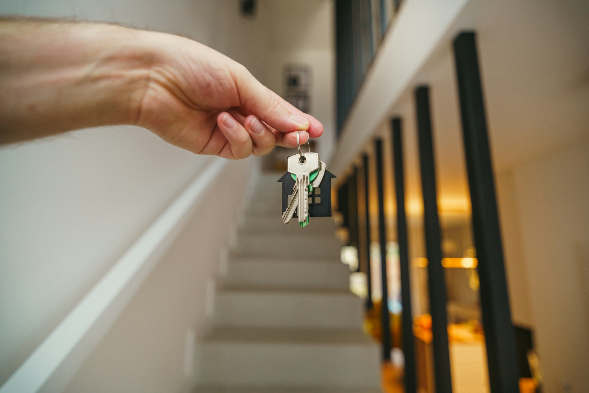 A hand holding a set of house keys on a house-shaped key ring at the foot of a white staircase