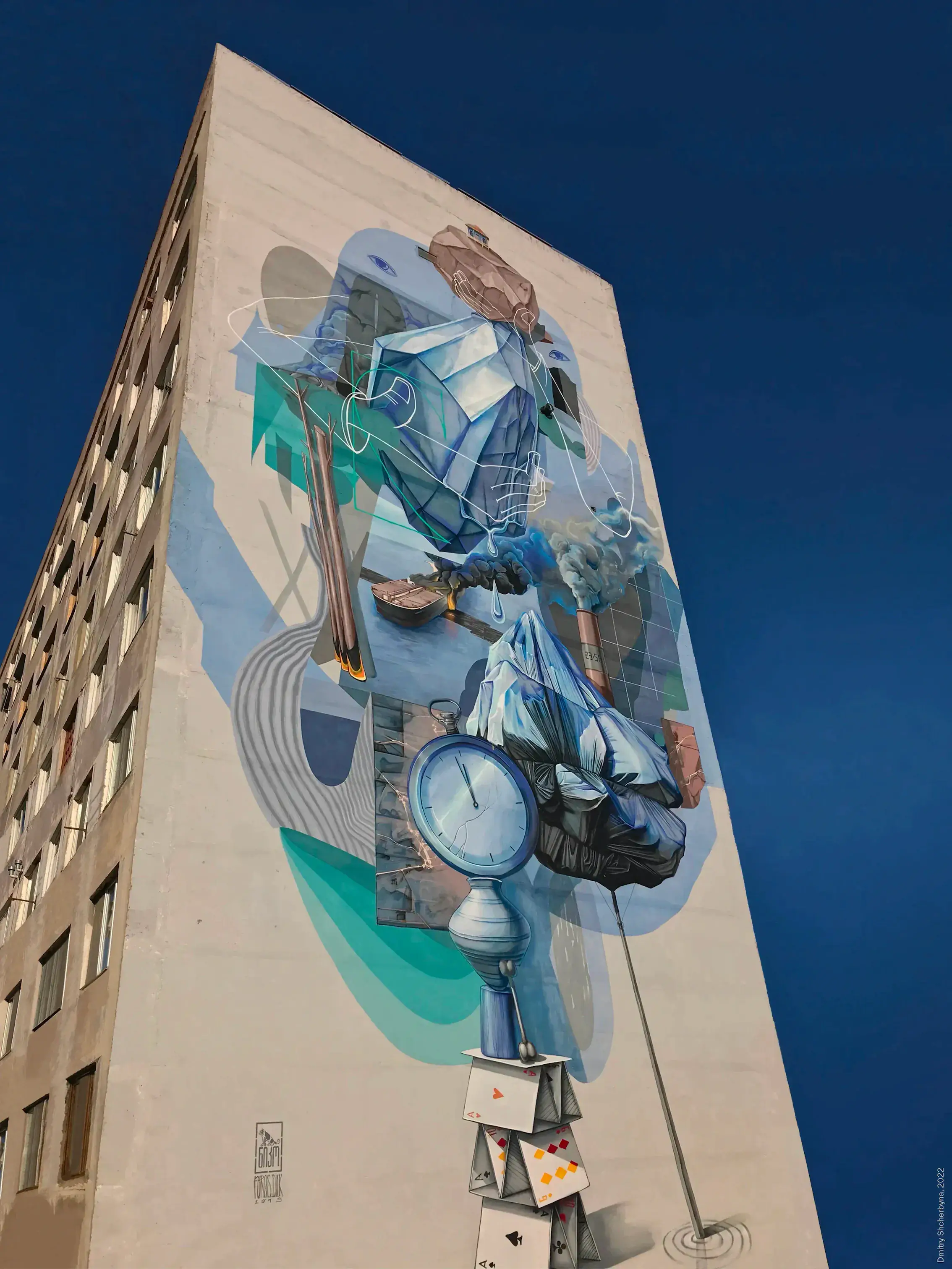 A tall surrealist mural on an apartment building consists of blue-toned objects stacked vertically - a suitcase, an ice block, a clock face, draped fabric, and a house of playing cards at the base - all connected by flowing lines against a deep blue sky.