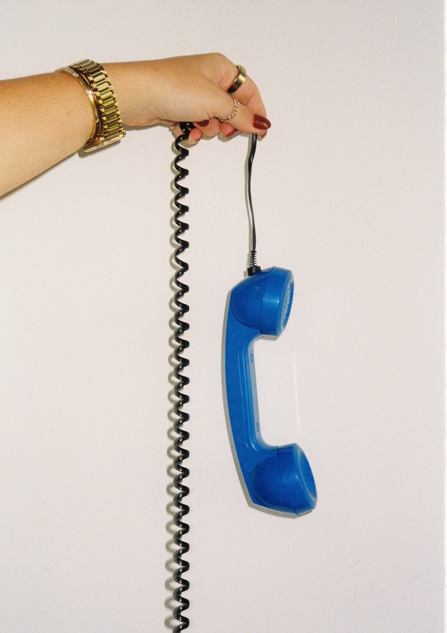 a hand with a gold watch holding up a blue corded phone in front of a white wall