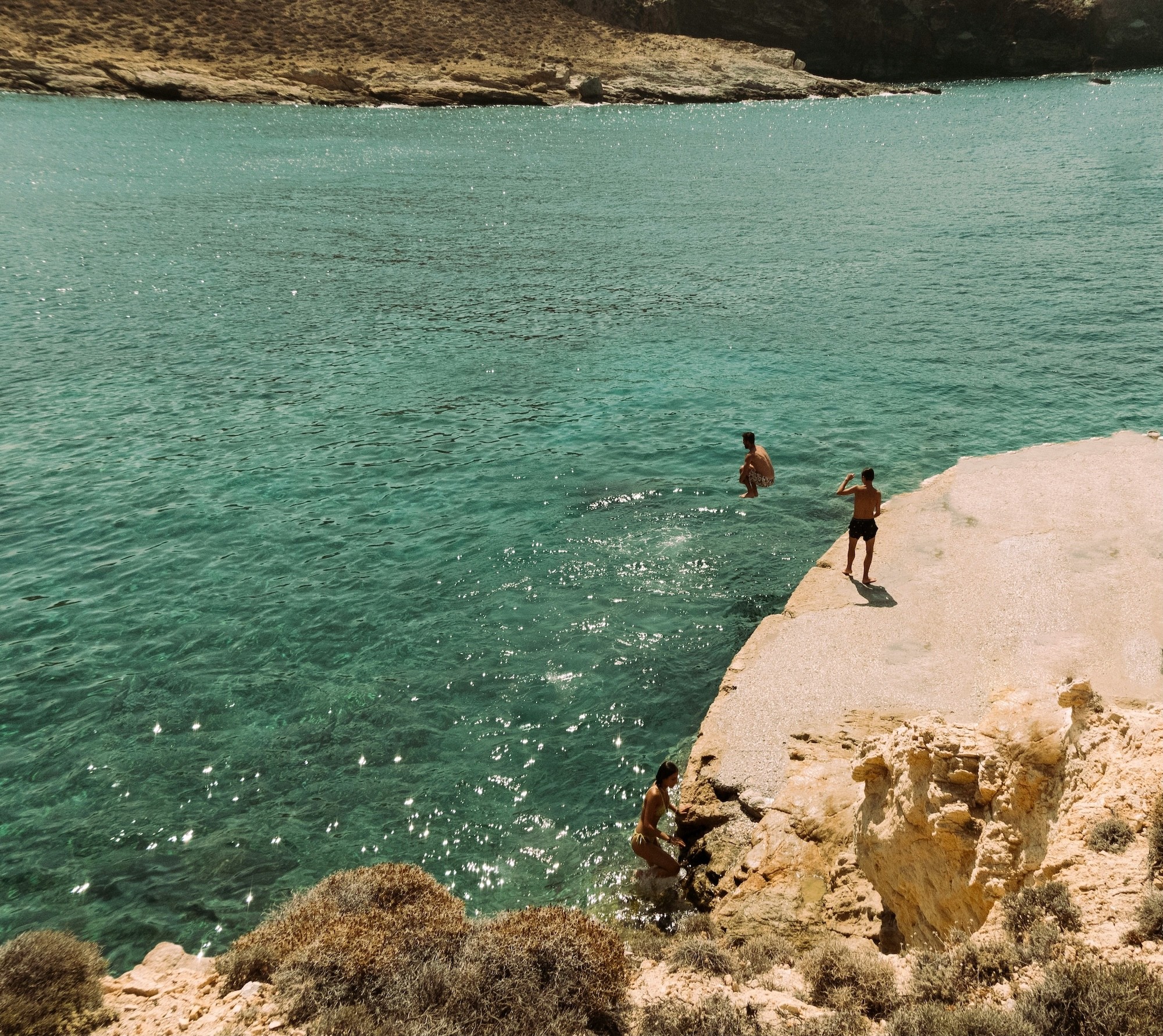 Swimmers diving into sparkling turquoise waters from sun-bleached rocks along a Greek island coastline