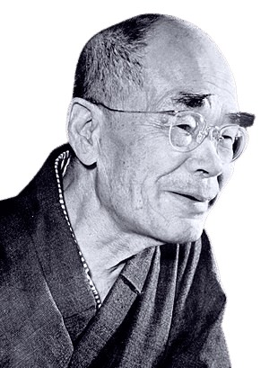 Black and white portrait of an elderly man with a shaved head, round glasses, and a gentle smile. He is dressed in traditional clothing with a visible patterned collar. His expression is serene and thoughtful, suggesting wisdom and a reflective demeanor.