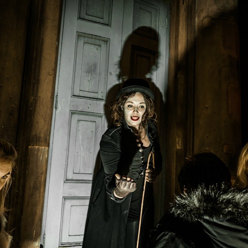 고딕 스타일의 의상을 입고 실크 해트를 쓴 여성이 문 앞에 서서 카메라를 향해 손을 내밀고 있습니다. 주변에는 몇몇 사람들이 보입니다.
