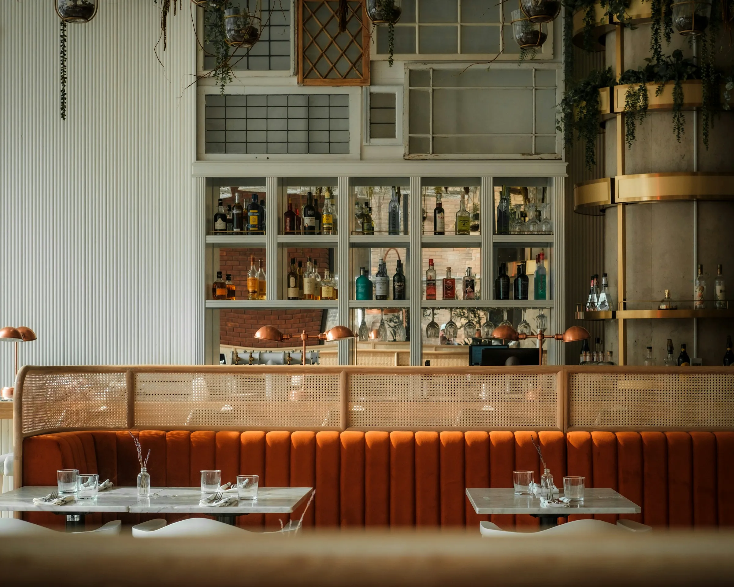 Restaurant interior with orange upholstered seating, marble tables, decorative glass shelving filled with bottles, and modern bar area design.