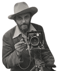 A black-and-white portrait of a bearded man wearing a wide-brimmed hat, holding a large-format camera outdoors. He is dressed in a casual suit jacket and shirt, appearing focused and ready to capture a photograph. The natural background of trees adds an earthy, adventurous feel, highlighting his connection to photography and the outdoors.