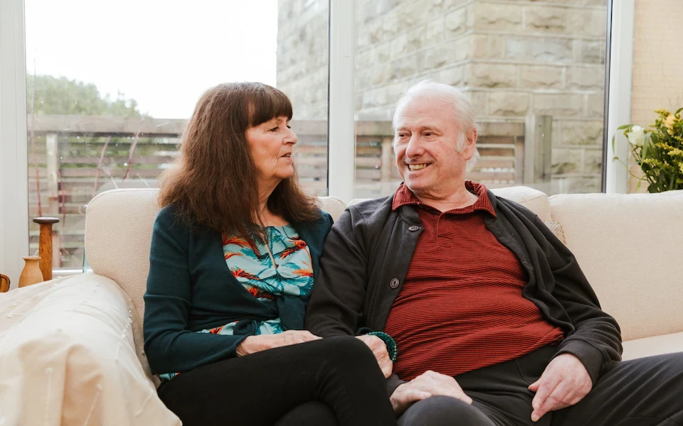 Older couple sitting on a sofa in a bright living room — Anne, an unpaid carer, supports her husband John.