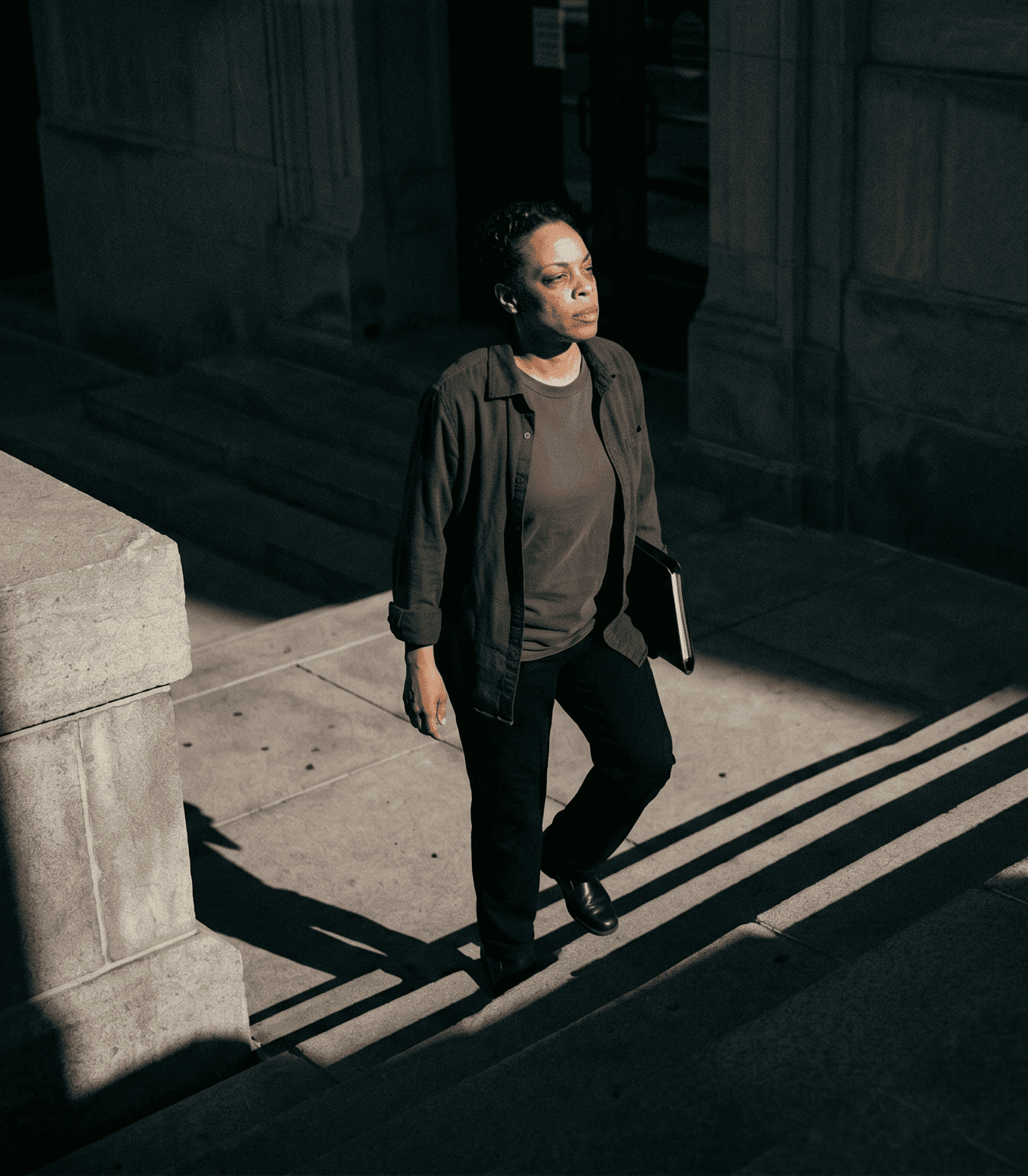 A person walks up a sunlit stone staircase, holding a closed laptop, with shadows cast across the steps.