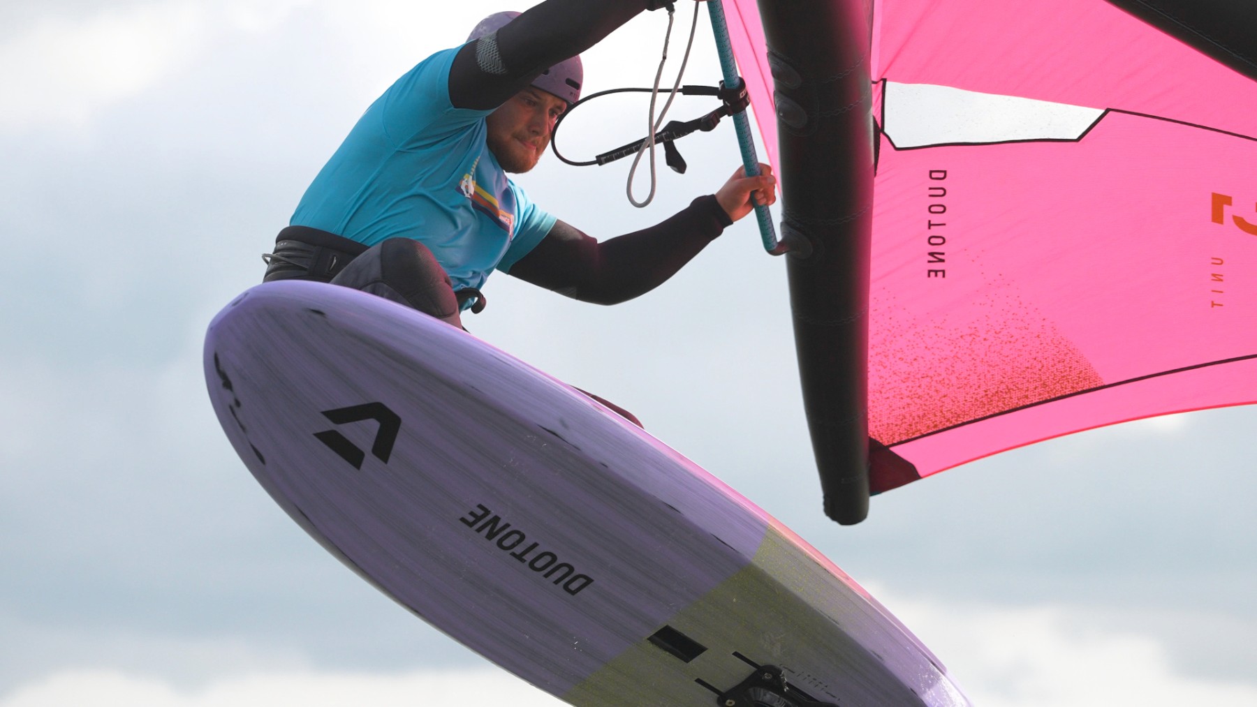 An instructor in a blue shirt and helmet soaring through the air on a purple and pink wing foil, with a cloudy sky backdrop, exuding excitement and focus.