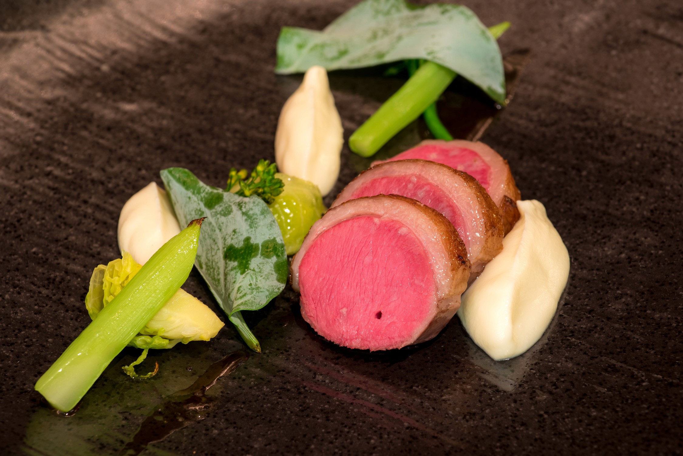 Slices of seared pink meat garnished with green herbs and white butter pats on a dark textured plate.