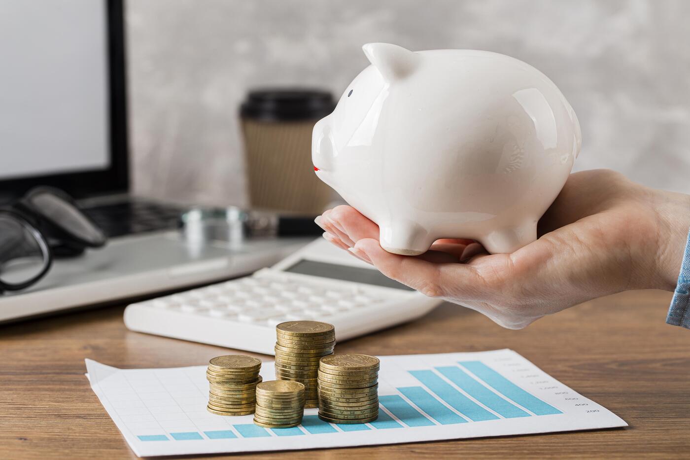 Foto de cofre em formato de porquinho sobre mesa com pilhas de moedas e planilha de crescimento, simbolizando controle financeiro e economia de custos administrativos