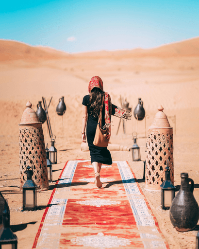 A person walks on a red carpet in a desert, flanked by decorative lanterns under a clear blue sky.