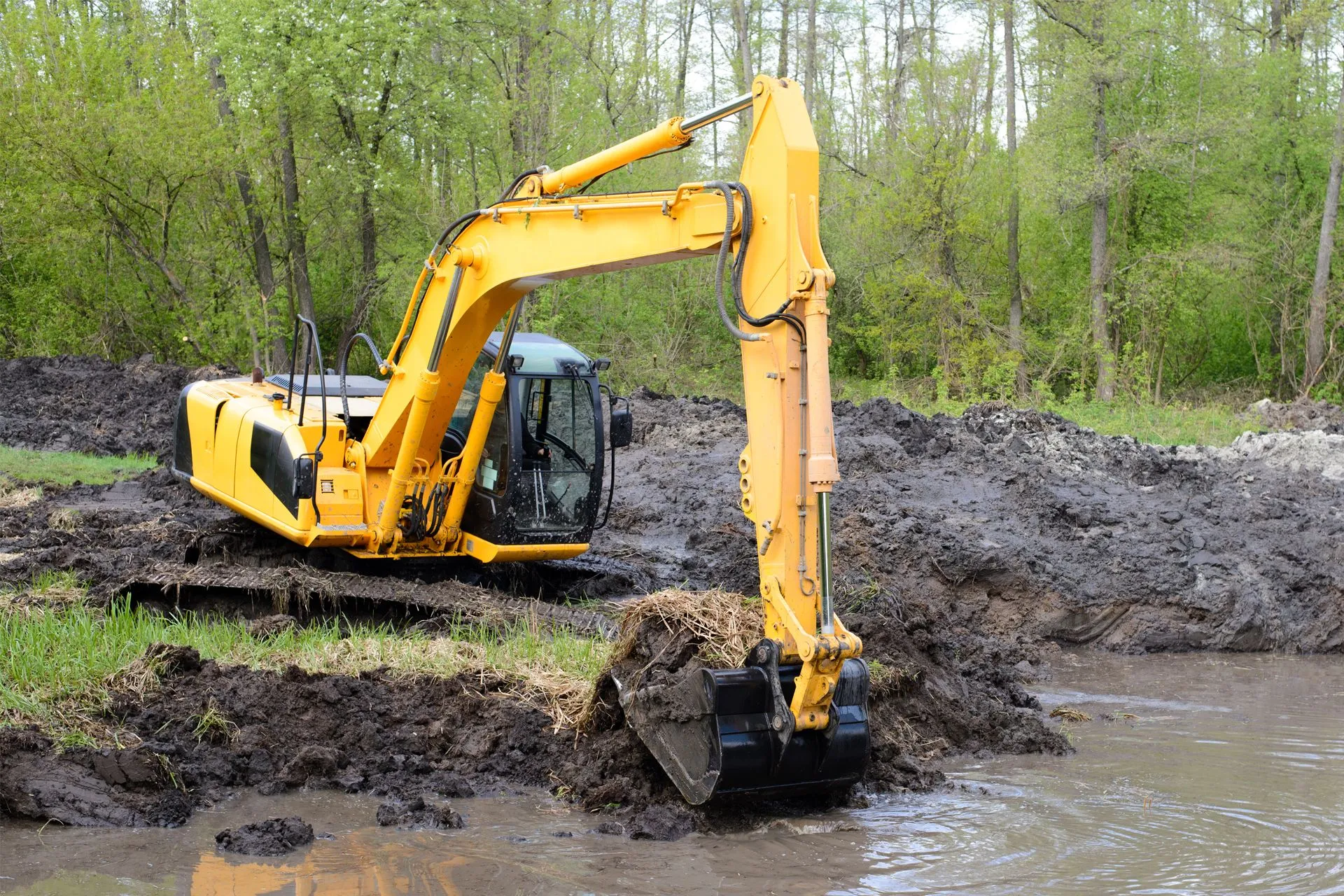 Excavator clearing mud and reshaping a wet, soft-ground area during site preparation near a wooded property.