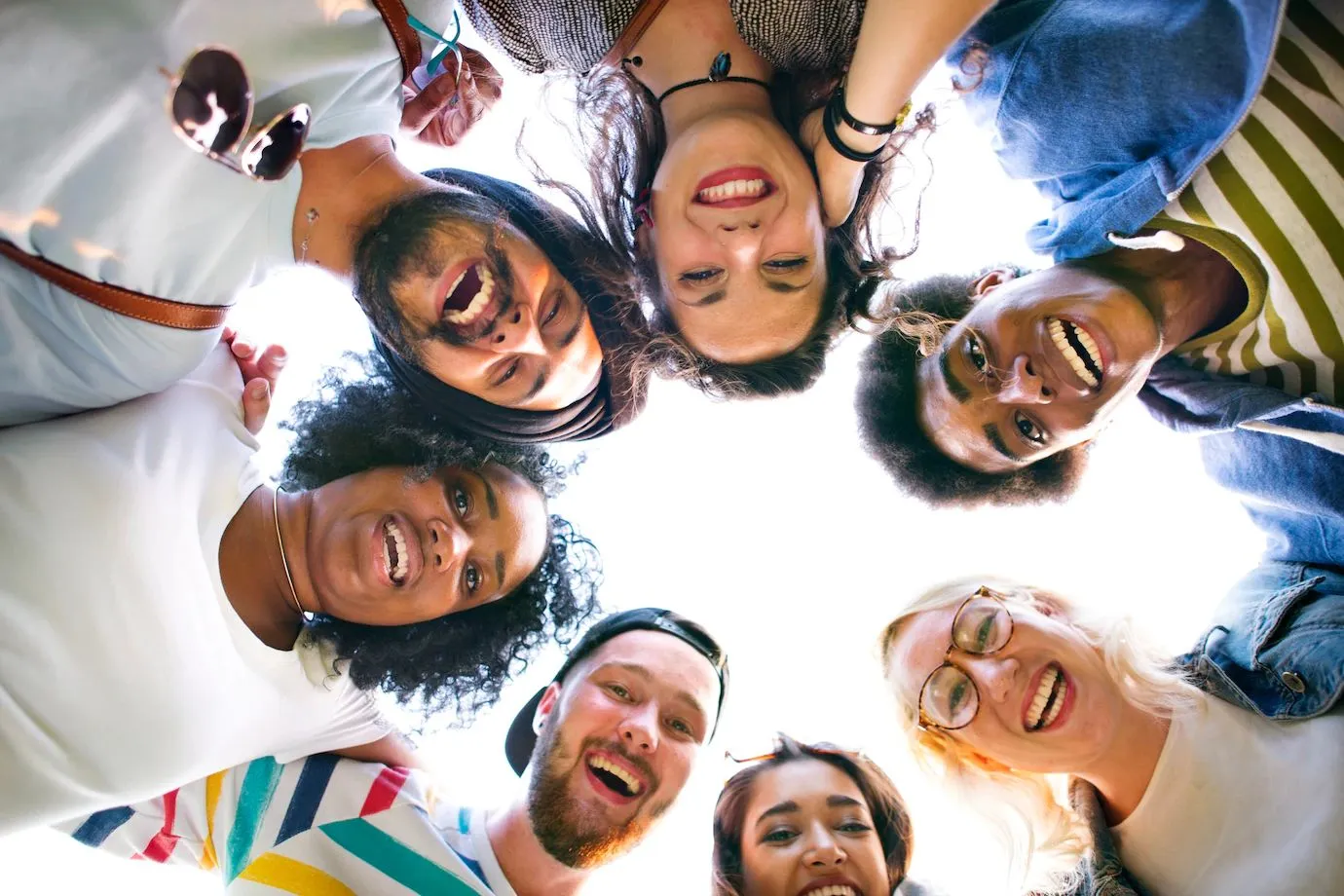 Um grupo diverso de pessoas forma um círculo e sorri para a câmera, criando uma imagem vibrante e alegre vista de baixo para cima. A foto destaca o espírito de união e diversidade, com diferentes etnias, estilos e expressões. A iluminação natural e as expressões de felicidade reforçam uma sensação de camaradagem e inclusão.