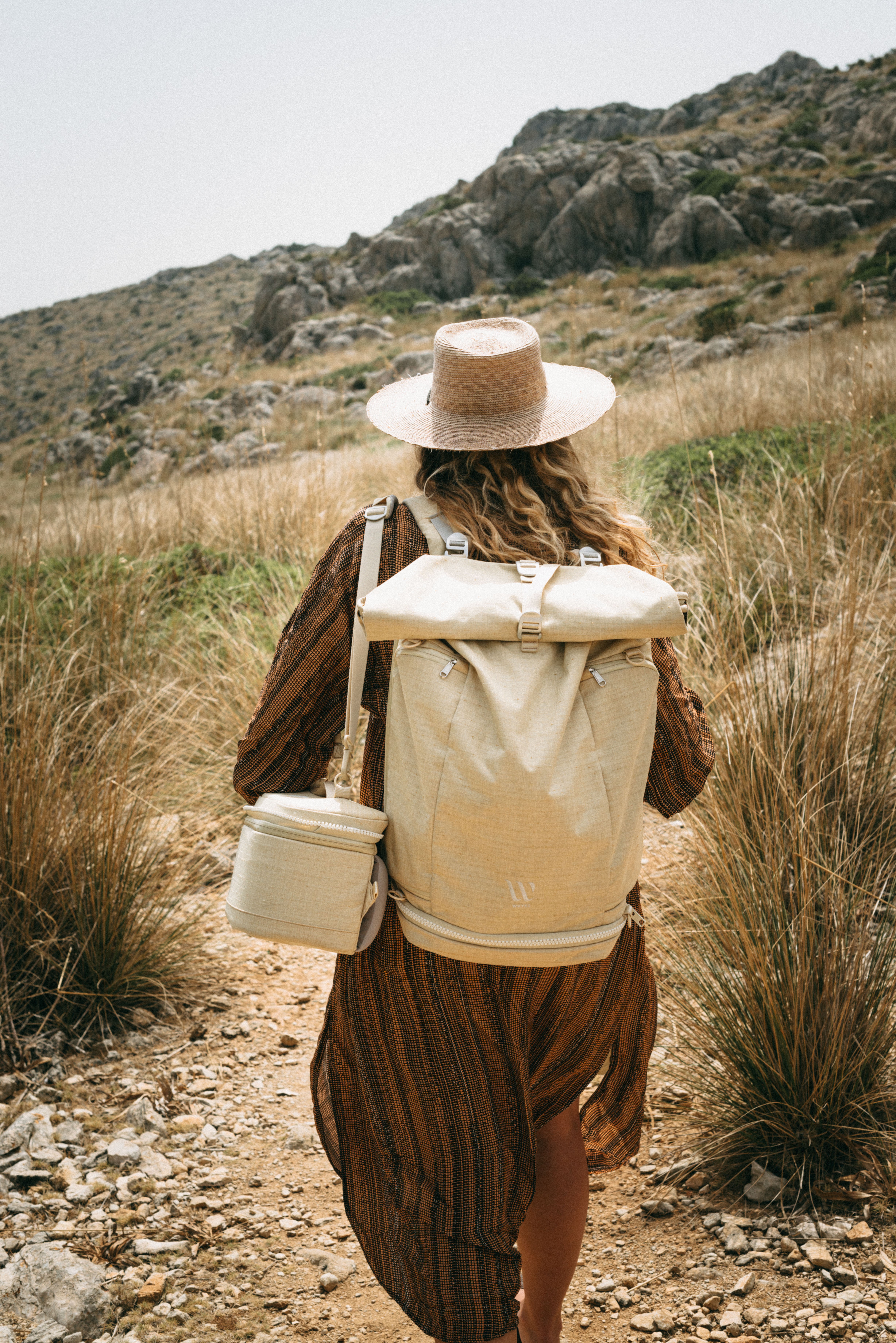 A woman in a flowing brown dress and straw hat walks along a rocky trail surrounded by tall grass and mountains, carrying a beige backpack and matching circular bag; keywords: outdoor adventure, hiking, nature exploration.