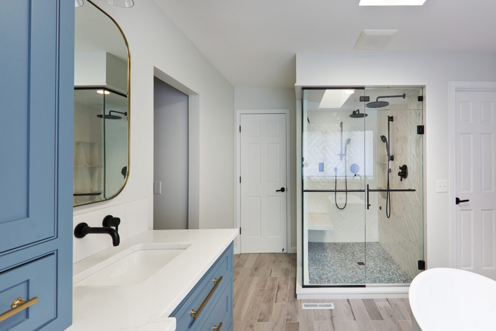 Bright modern bathroom featuring a white freestanding tub, light wood floors, large skylight, and built-in wooden and blue cabinetry. A walk-in glass shower with black fixtures sits near a private toilet room.