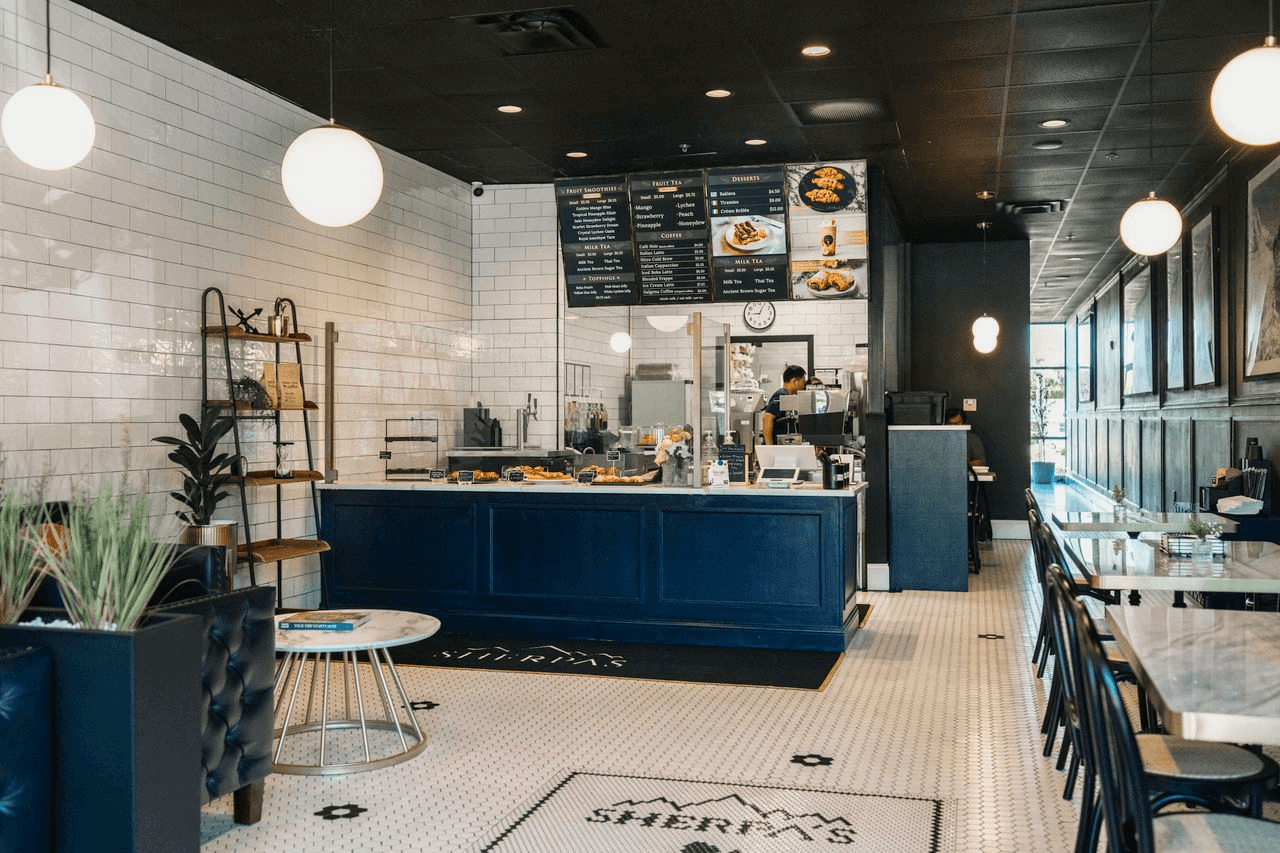 Inside Café Sherpa's in Brookfield, WI showing the navy blue service counter, overhead menu boards, barista at work, pendant lighting, and dining seating along the window. A welcoming café serving coffee, boba, tea, smoothies, and freshly baked pastries made in-house daily.