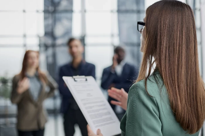 A imagem mostra uma mulher de costas, segurando uma prancheta com documentos, provavelmente em um ambiente corporativo. Ela parece estar apresentando ou explicando algo para um grupo de pessoas que estão à sua frente, desfocadas no fundo. As pessoas à frente estão usando trajes formais, sugerindo um ambiente de trabalho ou uma reunião de negócios. A cena parece retratar uma situação de introdução ou apresentação formal, possivelmente um processo de onboarding de novos funcionários ou uma reunião de equipe. O ambiente parece ser moderno, com grandes janelas ao fundo que deixam entrar muita luz natural.