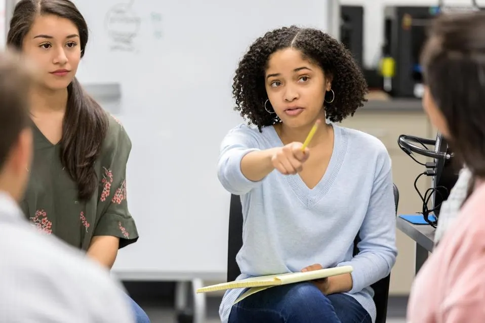 High school student holding a pencil and notebook actively debating with peers seated in a circle, symbolizing thoughtful discussion and student-led ethical inquiry.